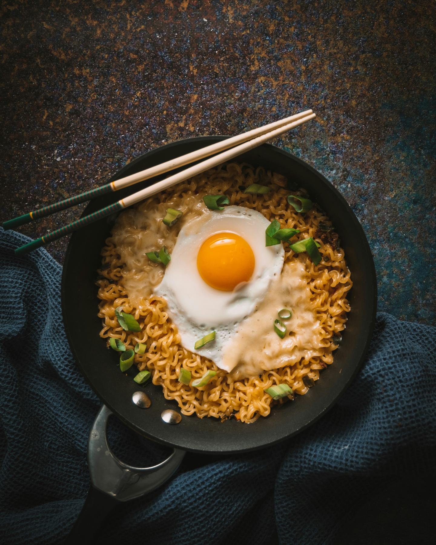 Instant noodles with a fried egg in a black pan on a dark surface, shot from above
