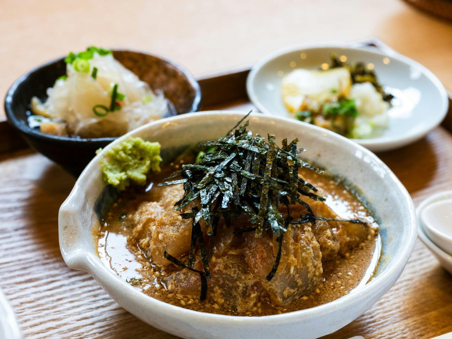 A Japanese rice bowl topped with shredded nori and broth, with small side dishes and chopsticks on a wooden table