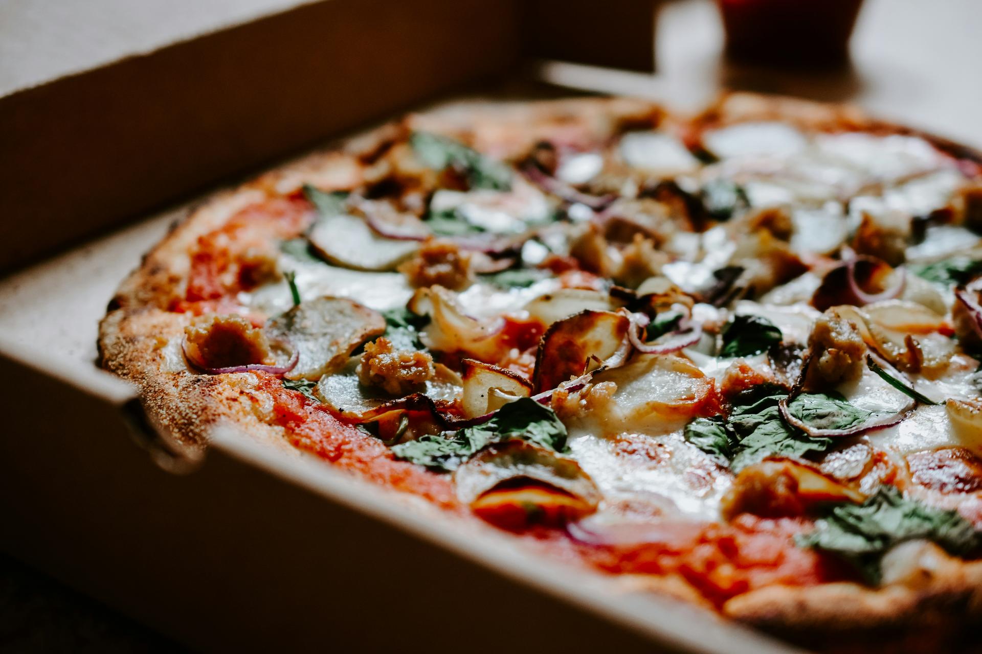 A pizza topped with tomato, mozzarella, sausage, red onion and spinach in an open cardboard delivery box
