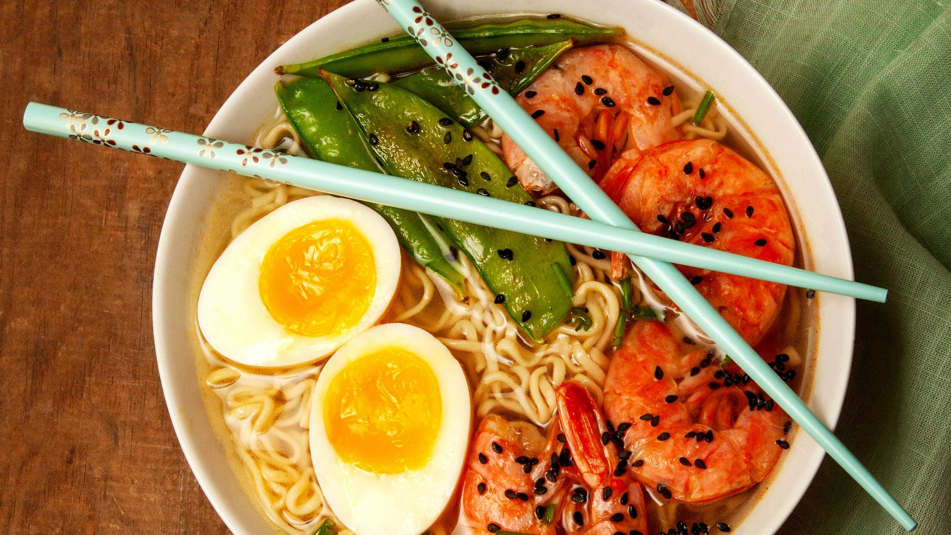 Overhead shot of a steaming bowl of ramen with soft-boiled eggs, shrimp and snap peas, chopsticks resting across the rim